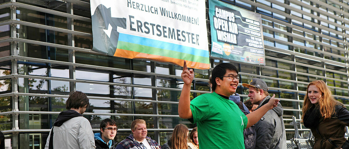 Ein Student in einem grüen T-Shirt steht vor der Hochchulbibliothek und zeigt mit seinen Händen in verschiedene Richtungen. Hinter ihm hängt ein großes Banner, auf dem die Erstsemester begrüßt werden.