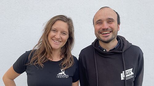 Carolin und Enrico stehen nebeneinander. Beide haben schwarze T-Shirts an. Auf Carolins steht "Robert Schmidt Institut" und auf Enricos "StartUpYard".