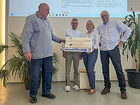 Die vier Personen stehen nebeneinander und halten den ca. 1 Meter breiten symbolischen Scheck in der Hand.