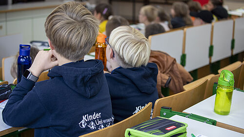 Es sind zwei Jungs von hinten zu sehen. Sie sitzen im Hörsaal und lauschen gespannt. In den REihen davor sind unscharf weitere Kinder zu erkennen.