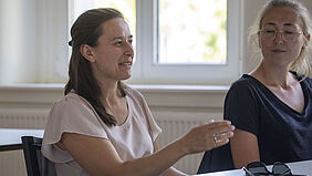 Two doctoral students are sitting in a seminar room. The woman on the left, wearing a light-coloured, short-sleeved jumper, is talking and gesticulating with her right hand. The woman on the right is wearing a black top, blonde hair and glasses and is listening.