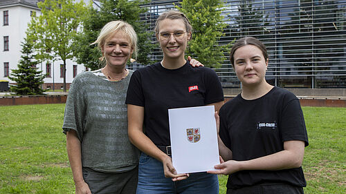 Die drei Personen stehen dicht nebeneinander, lächeln und halten die A4-Mappe mit dem Bescheid in den Händen. Im Hintergrund sind die Campuswiese und die Hochschulbibliothek zu sehen. 