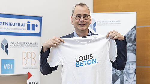 Der Professor hält vor seinem Körper ausgebreitet ein weißes T-Shirt in der Hand, auf dem steht "Louise Beton."