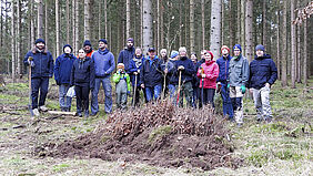 Die Gruppe steht hinter den Bündeln mit den knapp kniehohen Pflänzchen im Wald.