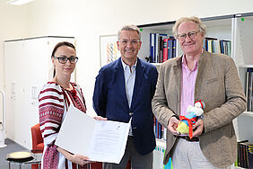 Fomenko Die drei Personen stehen lächelnd im Büro des Rektors. Dozentin Anna Fomenko präsentiert die Absichtserklärung und der Rektor hält die Puppe in beiden Händen.