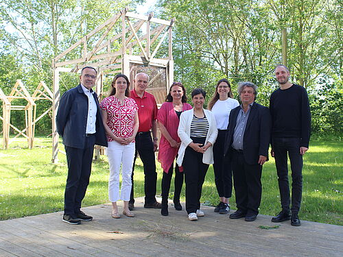 The picture shows a group of eight people standing outside on a wooden deck. In the background are trees and a wooden frame construction that looks like an unfinished building. The people stand next to each other and look into the camera. They are dressed in different casual and business attire. The sky is clear and it is a sunny day. The scenery looks friendly and relaxed.