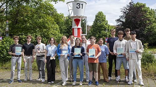 Die Gruppe steht draußen vor einem Seezeichen, einer rot-weiß-gestreiften Tonne, und hält Urkunden sowie Gutscheine in den Händen. Tonne