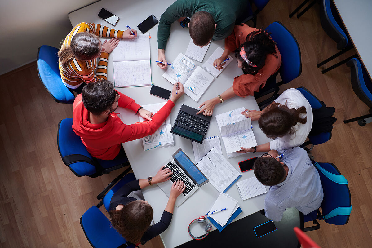 Introduction_Image_Symbolbild_WR_Master_SL_2023 Introduction_Image Studiengang Wirtschaftsrecht Master Studierende sitzen um einen Tisch herum auf dem Unterlagen verstreut sind, Draufsicht