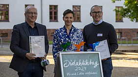 Die drei Personen stehen nebeneinander hinter einem Aufsteller mit dem Titel "Didaktiktag 2024". Sie halten ihre Urkunden in der Hand. Auf dem Aufsteller stehen die drei Preispokale in Form des Hochschulfischers.