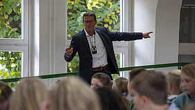 Der MAnn ist über die Köpfe einiger Kinder hinweg zu sehen. Er steht am Rande des Hörsaals und zeigt auf ein FEnster. 