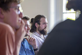 Drei junge Promovierende sitzen nebeneinander in einem Seminarraum und hören aufmerksam zu. Der Mann in der Mitte trägt einen Vollbart und hat lange Haare, die zu einem Knoten gebunden sind. Die anderen beiden Männer tragen Brillen und haben den Blick nach vorne gerichtet.
