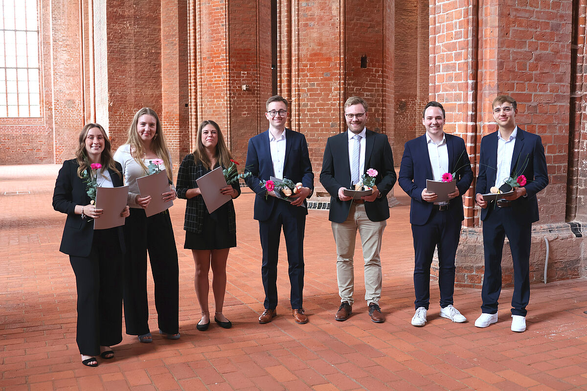 Auf dem Foto sind die Absolventen des Bachelor Studiengangs Wirtschaftsrecht zu sehen. Alle in feierlicher Kleidung. Im Hintergrund links ein großes Kirchenfenster durch das die Sonne hereinstrahlt. Der Innenraum der St. Georgenkirche besteht aus leuchtend rotem Backstein.