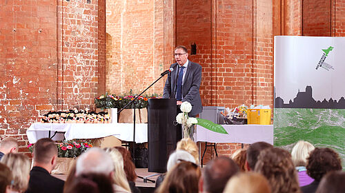 Der Dekan der Fakultät Wirtschaftswissenschaft, Prof. Dr. Kai Neumann auf der Bühne vor dem Rednerpult. Im Hintergrund der Aufsteller der Fakultät Wirtschaftswissenschaften in weiß mit grünem Logo. Im Vordergrund ist das sitzende Publikum zu erahnen.