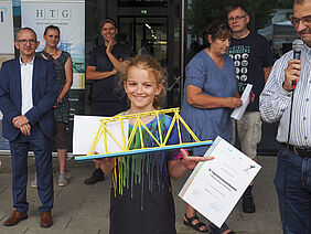 Sigrun hält lächelnd ihre Urkunde und die Papierbrücke in den Händen. Sie steht vor der Glasfront des Einganges zum Haus 6.