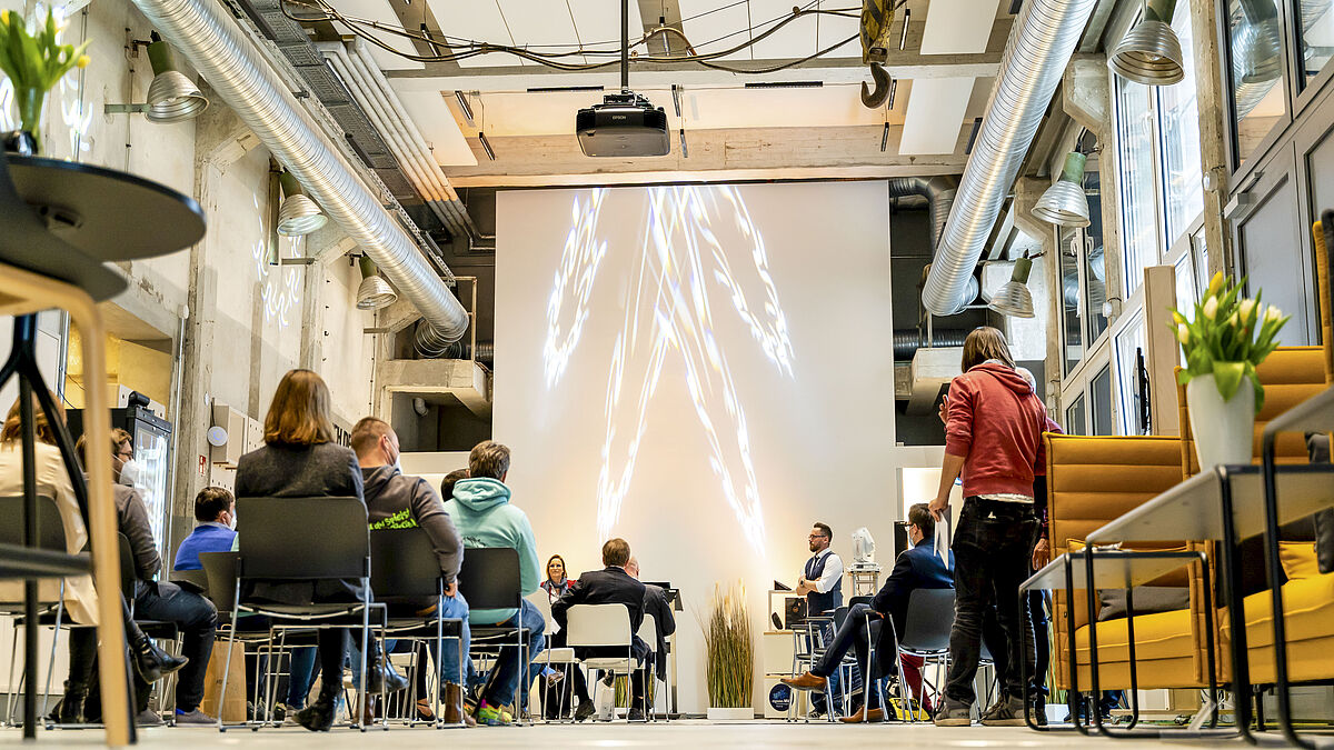 InnovationPort Wismar Eine Person steht vor einer Präsentation. Zuschauer, die auf Stühlen sitzen, sind von hinten zu sehen.