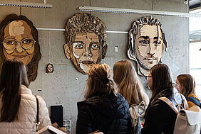 Es sind Schülerinnen von hinten zu sehen, die überlebensgroße Gesichter betrachten. Diese Gesichter hängen an einer Sichtbetonwand.