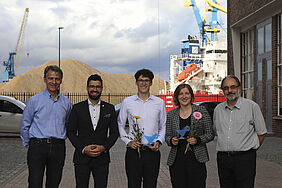 Die fünf Personen stehen nebeneinander. Im Hintergrund ist ein Schiff, ein Kran und ein Kieshaufen im Wismarer Seehafen zu erkennen.