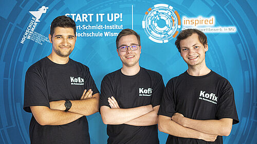 Die drei jungen Männer sind mit schwarz T-Shirts bekleidet, auf denen "KOFIX die Rucksackköche" steht. Sie stehen vor einer hellblauen Wand, auf der zwischen dem Logo der Hochschule Wismar und dem Logo des Landesideenwettbewerbes "Start it up! Beim Robert-Schmidt-Institut der Hochschule Wismar" steht.