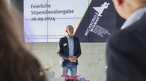 Der Fotograf schaut über die Schulter zweier Stipendiaten. Der Prorektor steht vor einer Bildschirmpräsentation auf der Datum und Veranstaltungstitel stehen.
