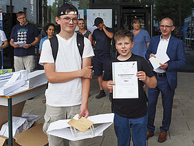 Die beiden Jungs halten ihre Urkunde und Papierbrücke in den Händen.  Beide stehen vor der Glasfront des Einganges zum Haus 6.