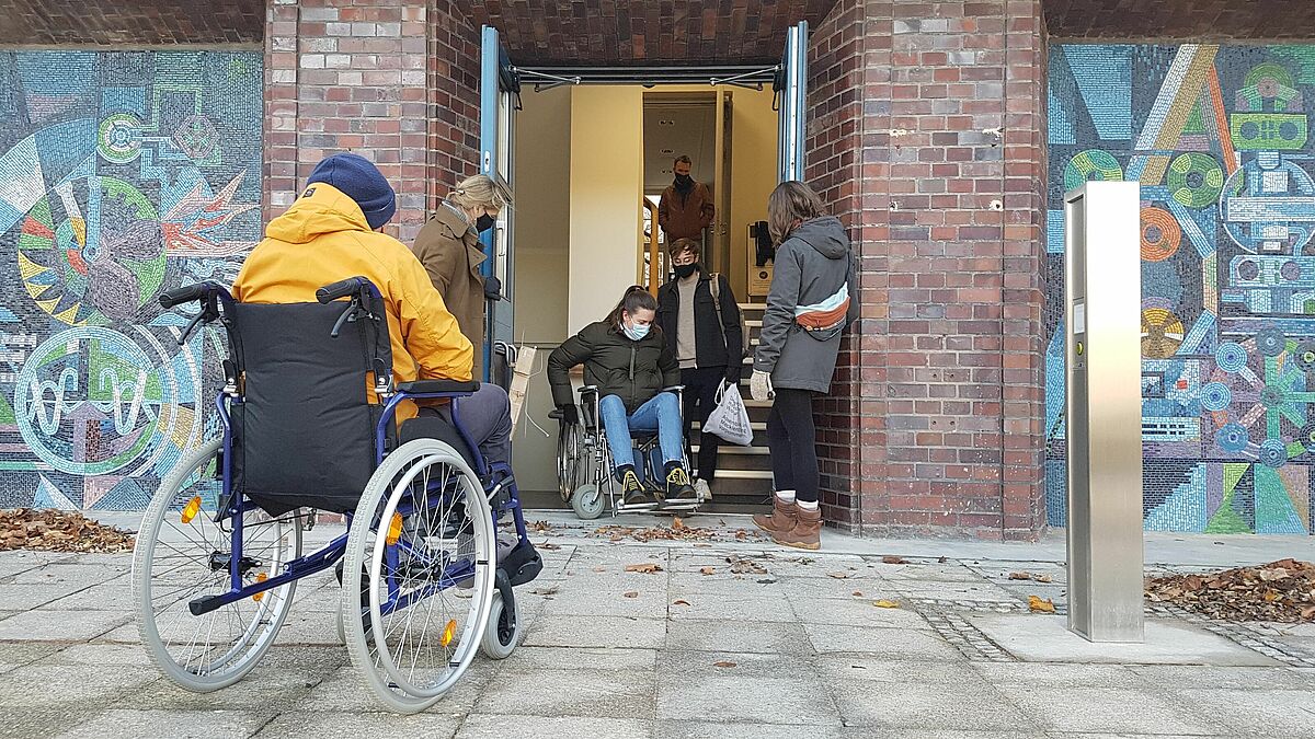 Es sind zwei Studierende im Rollatuhl am Eingang des Hauses 18 auf dem Wismarer Campus zu sehen.