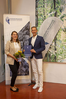 Die beiden Persoenen stehen vor einem Banner mit dem Aufdruck "Ingenieurkammer M-V". Die Ausgezeichnete hält Blumen und Urkunde in der Hand.