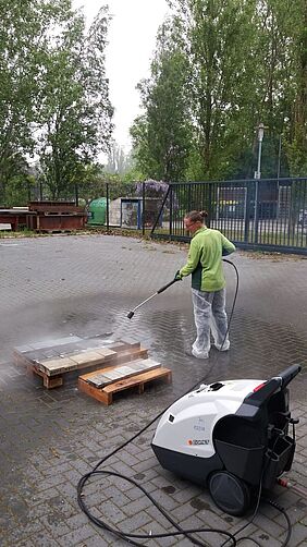 Sandra Jäntsch seht mit Schutzhosen und -handschuhen bekleidet hinter BEtonplatten, die auf Holzgestellen liegen und hat einen Hochdruckreiniger in der Hand.