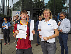 Felix hält seine Urkunde und Papierbrücke in den Händen, die Frau einen Umschlag mit dem Preisgeld. Beide stehen vor der Glasfront des Einganges zum Haus 6.