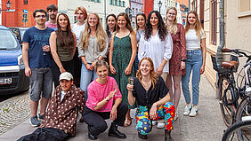 Die Personen stehen als Gruppe auf dem Fußweg. Ein Student liegt schräg aufgestützt und zwei Studentinnen prosten sich zu. Im Hintergrund ist eine typisch hanseatische Häuserzeile zu sehen.