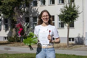 Die Preisträgerin steht auf der Wiese vor einem Hochschulgebäude und hält lachend ihre Urkunde sowie den Blumenstrauß in der Hand.