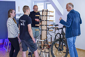 An einem Holzständer sind übereinander fünf Fahrradlenkerr angebracht. Diese sind mit unterschiedlichen Holzgriffen bestückt. Um diesen Ständer herum stehen vier Personen und unterhalten sich.