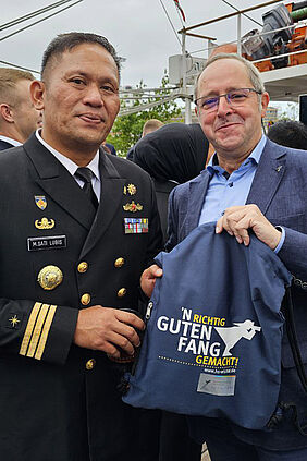 Die beiden Männer schauen in die Kamera und Präsentieren einen Beutel mit dem Hochschulfischer und der Aufschrift "N´richtig guten Fang gemacht"."