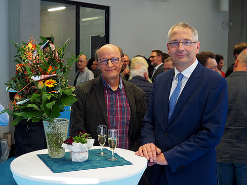 Die beiden genannten Personen stehen an einem Tisch, auf dem sich zwei Sektgläser und ein großer Blumenstrauß befinden. Im Hintergrund sind zahlreiche Gäste zu sehen.