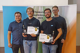 Lukas und Leif stehen gemeinsam mit zwei Ausgezeichneten, diese mit BLumen und Urkunden in der Hand, gemeinsam vor einer Bannerwand, auf der Feierliche Immatrikulation 2023 steht