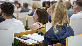 Der Blick wird über die Schulter einer Teilnehmerin auf weiter gelenkt, wobei Block und Taschenrechner im Fokus stehen.